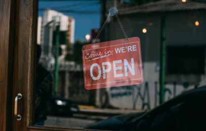 Bildausschnitt: Glastür eines Geschäfts, in dem sich die Stadt spiegelt, mit rotem Geöffnet Schild; Come in we're open